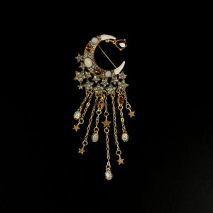 May include: A celestial-themed brooch featuring a crescent moon design with white enamel tips and adorned with stars, rhinestones, and dangling chains with star and teardrop charms. The brooch has a gold-tone finish.