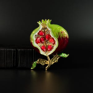 May include: A pomegranate brooch featuring a gold-tone metal stem and leaves accented with small green crystals. The fruit is rendered in deep red and bright green enamel, with the interior showing red crystal seeds set against a white background.