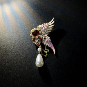 May include: A gold-toned brooch with a wing design, featuring pink and white enamel, a large red gemstone, and small red and white accents. A dangling white teardrop pearl hangs from a chain. The brooch is set against a dark background.