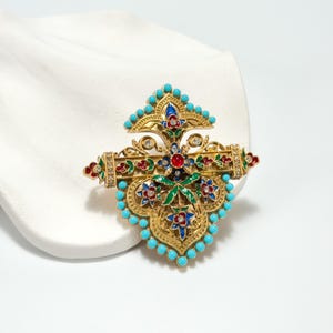 May include: Ornate gold-tone brooch with turquoise, red, blue, and green enamel details. The brooch features a central design with floral and jewel accents, surrounded by turquoise beads. A decorative piece of jewelry.