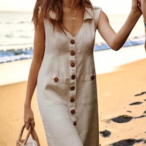 Może przedstawiać: Beżowa sukienka bez rękawów z guzikami. Sukienka ma dekolt w serek, dwie przednie kieszenie i brązowe guziki. Pleciona torba z koszyka jest trzymana w lewej ręce. Sukienka wydaje się być wykonana z lnu.