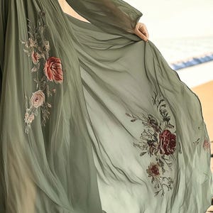 May include: A flowing, olive green dress with long sleeves and floral embroidery. The sheer fabric drapes elegantly, revealing intricate details of red and pink roses and other flowers. The dress is a romantic and ethereal garment.