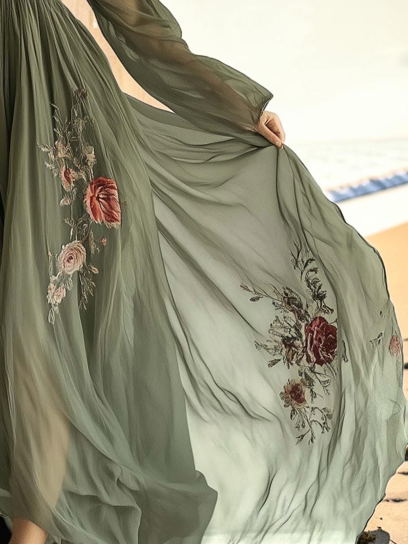 May include: A flowing, olive green dress with long sleeves and floral embroidery. The sheer fabric drapes elegantly, revealing intricate details of red and pink roses and other flowers. The dress is a romantic and ethereal garment.