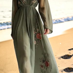 May include: A flowing, olive green maxi dress with long sleeves and a deep V-neck. The dress features floral embroidery on the bodice and skirt. The sheer fabric creates a light and airy look, perfect for a beach setting.