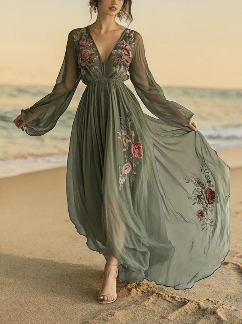 May include: A flowing, olive green maxi dress with long sleeves and a deep V-neck. The dress features floral embroidery on the bodice and skirt. The model is walking on a sandy beach, with the ocean in the background.