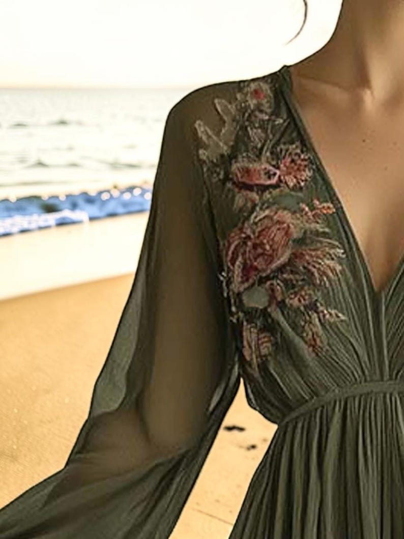 May include: Olive green dress with long, sheer sleeves and a deep V-neck. The dress features floral embroidery in shades of red, pink, and green on the shoulder. The dress is flowing and appears to be made of a lightweight fabric.