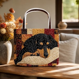 May include: A quilted tote bag featuring a black and tan dachshund puppy sleeping on a cream-colored pillow. The bag has a patchwork background of warm brown, orange, and red squares with white handles and intricate wavy stitching throughout.