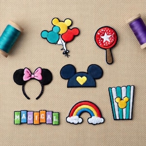 Könnte beinhalten: Gestickte Aufnäher mit Designs von Zeichentrickfiguren. Enthält ein Minnie Mouse-Stirnband, Mickey Mouse-Ohren, einen Regenbogen, Popcorn, Luftballons und einen Lutscher. Das Wort "MAGICAL" ist ebenfalls auf einzelnen Aufnähern geschrieben.