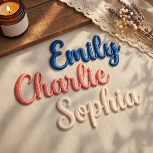 Puede incluir: Parches de nombres bordados en escritura azul, coral y crema. Se muestran los nombres "Emily", "Charlie" y "Sophia". Los parches están sobre una tela color crema. Una vela encendida y flores secas están en el fondo.