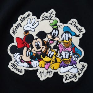 Peut inclure: Écusson brodé représentant Mickey Mouse, Minnie Mouse, Dingo, Donald Duck, Daisy Duck et Pluto. L'écusson a un fond crème avec les personnages dans leurs couleurs classiques. Les noms des personnages sont brodés autour d'eux.