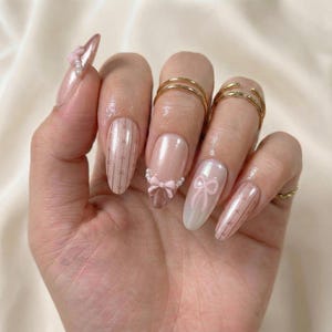 May include: A hand with long, almond-shaped nails. The nails are painted in a light pink hue with various designs, including a bow, pearl accents, and striped patterns. Gold rings adorn the fingers.