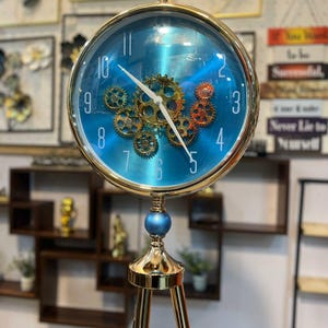 May include: A gold-coloured, steampunk-style clock with a blue face and visible gears. The clock has a round face with white numerals and hands. It stands on a tripod base with black legs and a blue sphere accent.