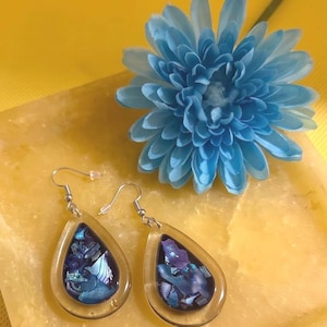 May include: A pair of teardrop-shaped earrings featuring a clear resin outer border and a dark blue interior filled with iridescent, multi-tonal blue and purple crushed foil fragments. The earrings hang from silver-toned metal fishhook wires.