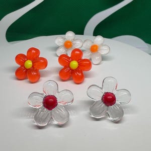 May include: Three pairs of flower-shaped stud earrings made of translucent material. One pair is orange with yellow centers, one is clear with orange centers, and one is clear with bright red centers, all featuring five rounded petals.