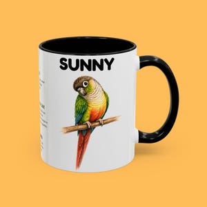 Könnte beinhalten: Weißer Keramikbecher mit schwarzem Innenraum und Griff. Der Becher zeigt eine farbenfrohe Illustration eines Papageis, der auf einem Ast sitzt, mit dem Wort "SUNNY" darüber. Der Papagei hat grüne, gelbe, rote und braune Federn.