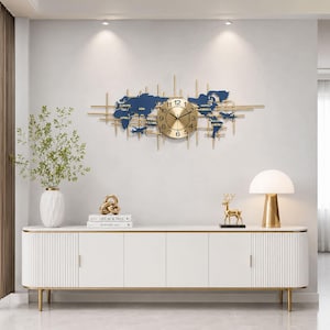 May include: A gold and blue world map wall clock. The clock features a gold-colored clock face with black hands and numbers. The map is blue with gold accents and includes the names of continents. The clock is mounted on a white wall above a white cabinet.