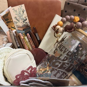 May include: A gift box containing a glass tumbler with a bamboo lid and the text NEVER TOO COLD FOR ICED COFFEE, a brown notebook, a cream-coloured pouch, a beaded keychain, pens, a bookmark with a book illustration, and a small white crochet hat.