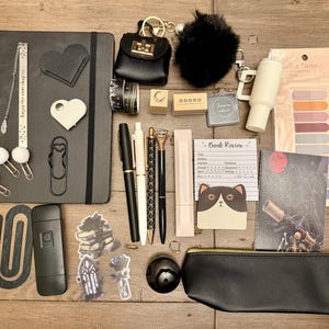May include: An assortment of stationery items, including a black journal, pens, stickers, and a pencil case, arranged on a wooden surface. Accessories include a keychain, pom-poms, and paper clips. The items are in various shades of black, white, and gold.