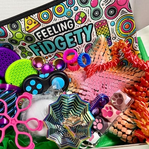 May include: A gift box filled with various colourful fidget toys, including a zippered pouch printed with the text FEELING FIDGETY, a purple and green textured disc, a black controller, a pink star-shaped ring, and a multi-coloured metal spinner.