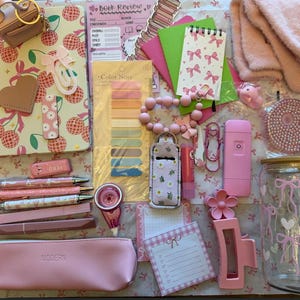 May include: A collection of stationery and accessories including a cherry-patterned notebook, a pink pencil case, pens, sticky notes, a glass cup with a bow print, a pink hair clip, fuzzy socks, a small portable fan, and various paper goods.