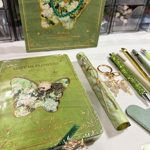 May include: A green stationery set featuring a notebook with the text LOST IN FLOWERS and a butterfly cutout, a rolled paper scroll tied with twine, a butterfly keychain, a green glitter pen, and a matching green gift bag with butterfly art.