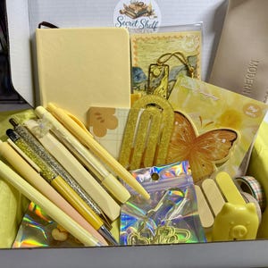 May include: A stationery gift box containing a yellow notebook, a beige pencil case with the word MODERN, a gold glitter arch bookmark, a butterfly-shaped notepad, yellow pens, a pig-shaped eraser, and various gold-toned paper clips in a pouch.