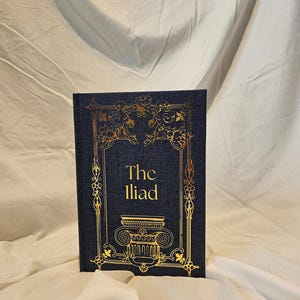 May include: A hardcover book titled The Iliad with a dark blue fabric cover. The front cover features gold foil stamping with an ornate rectangular border, a central column capital illustration, and the text The Iliad in gold lettering.