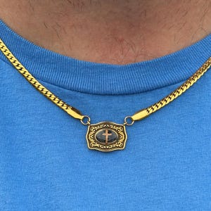 May include: Gold-toned necklace featuring a rectangular pendant with a cross design. The pendant is attached to a flat, herringbone chain. The necklace is worn against a blue shirt.