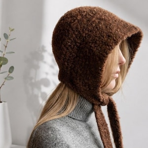May include: A dark brown bonnet crafted from a textured, fuzzy knit material. The headpiece features a rounded crown that covers the head and ears, with two long, matching knit ties extending from the chin area to secure the garment in place.