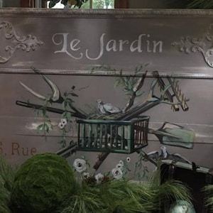 May include: A painted wooden sign with the words "Le Jardin" and "6. Rue" in white lettering. The sign features a bird in a cage, gardening tools, and a green moss ball.