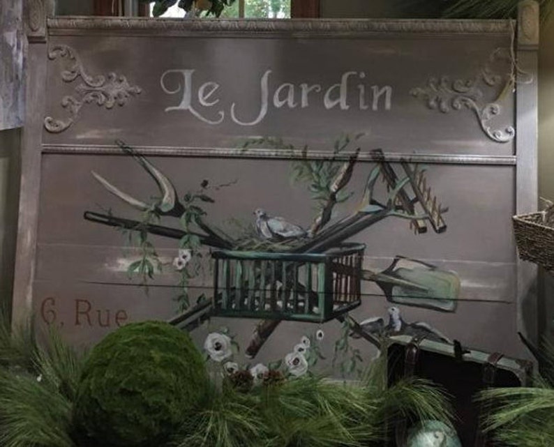 May include: A painted wooden sign with the words "Le Jardin" and "6. Rue" in white lettering. The sign features a bird in a cage, gardening tools, and a green moss ball.