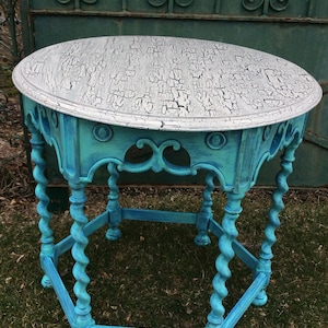 May include: A turquoise blue round table with a white cracked paint finish on the top. The table has a hexagonal base with twisted legs.