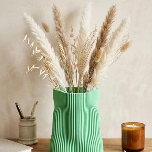 Puede incluir: Un jarrón verde menta texturizado, lleno de hierbas ornamentales secas. El jarrón tiene un diseño ondulado único. Una vela encendida en un tarro de cristal y un pequeño portalápices de cerámica también están sobre la superficie de madera.