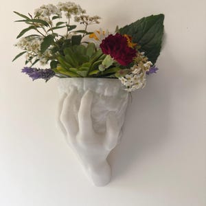 Può includere: Un vaso da parete bianco a forma di mano riempito con una varietà di fiori e verde colorati. La composizione comprende fiori bianchi, rossi e gialli, insieme a foglie verdi e piante grasse. Il vaso è montato su una parete bianca.