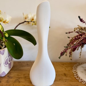 May include: A tall, white vase with a textured, ribbed design. The vase has a slender, elongated shape with a flared base and a curved top. The vase is displayed on a wooden surface, with plants in the background.