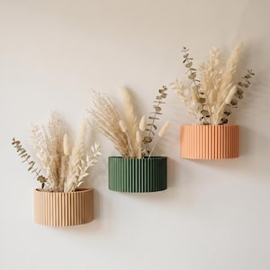 Puede incluir: Tres jardineras murales en beige, verde y melocotón, cada una con un diseño acanalado. Las jardineras contienen hierbas ornamentales secas y ramas de eucalipto, creando una estética minimalista y natural. Las jardineras están montadas en una pared de color neutro.