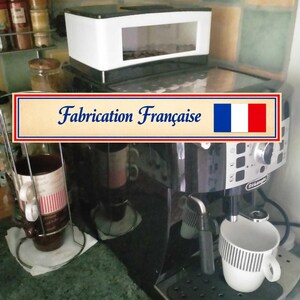 Op de afbeelding: Een zwarte DeLonghi espressomachine met een witte bonenreservoir bovenop en een witte mok met een zwart ruitpatroon op de lekbak. Een rechthoekig bord met een blauwe, witte en rode vlag bevat de tekst "Fabrication Française" in blauw.