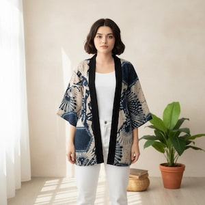 Puede incluir: Chaqueta estilo kimono con estampado azul marino y beige, ribete negro y mangas cortas. La persona lleva un top y pantalones blancos. Una planta verde en maceta se ve en el fondo.