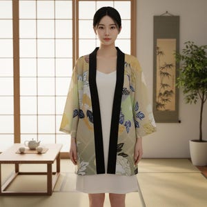 Op de afbeelding: Een vrouw draagt een beige kimono met een bloemenpatroon en zwarte bies. De kimono heeft een witte voering en wordt over een witte jurk gedragen. De achtergrond toont een traditioneel Japans interieur met een theetafel en een rol.