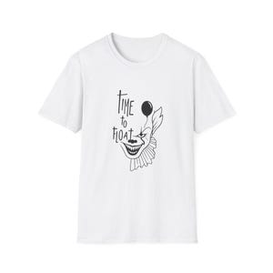 Può includere: T-shirt bianca con grafica in bianco e nero di una faccia da clown e la scritta "Time to Float". Il clown ha un sorriso sinistro e un palloncino. La maglietta è girocollo.