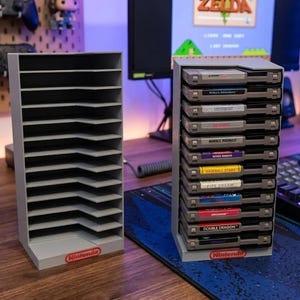 Puede incluir: Dos soportes de almacenamiento verticales de plástico gris para cartuchos de juego Nintendo. Un soporte está vacío y el otro está lleno con diez cartuchos grises. Cada soporte tiene un logotipo rojo de Nintendo en la base y diez ranuras.