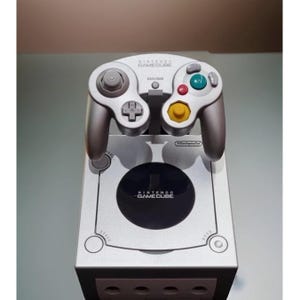 May include: Silver Nintendo GameCube console with a matching controller. The controller has grey, red, green, and yellow buttons. The console and controller are displayed on a light-coloured surface.