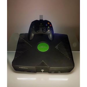 May include: Black Xbox console with a matching black controller. The console has a green circular button and the word "XBOX" printed on the front. The controller is placed on top of the console.