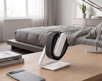 Pode incluir: Um suporte de carregamento branco segura um smartwatch preto com uma pulseira de tecido texturizado. O suporte possui um braço de apoio curvo e uma base plana. Um smartphone cinza exibindo a hora 7:15 repousa sobre a superfície de madeira.