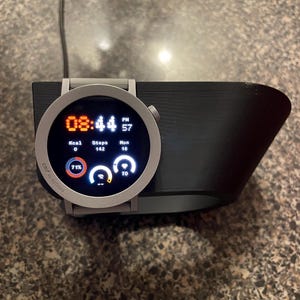 Puede incluir: Un reloj inteligente plateado con una pantalla negra que muestra la hora, los pasos y otros datos, apoyado en una base de carga negra. La esfera del reloj muestra la hora como 20:44. La base está sobre una superficie marrón moteada.