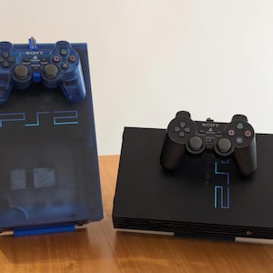 May include: A translucent blue PlayStation 2 console with a matching blue controller and a black PlayStation 2 console with a black controller. Both consoles feature the PS2 logo on the front, with the controllers resting on top of each unit.