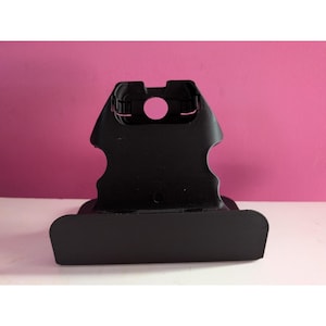 Op de afbeelding: Een zwart plastic object met een gebogen basis en een bovenste gedeelte met een cirkelvormig gat. Het object heeft een matte afwerking en staat tegen een roze achtergrond.