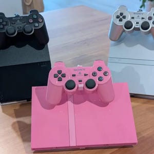 May include: Three Sony PlayStation 2 game consoles with matching controllers. One console and controller are black, one set is silver, and one set is pink. Each console is positioned vertically or horizontally with its controller resting on top.