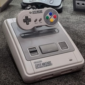 Peut inclure: Une console de jeu Super Nintendo Entertainment System gris clair avec une manette assortie posée dessus. La console comprend un interrupteur d'alimentation, un bouton d'éjection, un bouton de réinitialisation et le logo Super Nintendo imprimé.