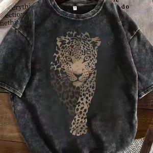 May include: A dark gray t-shirt featuring a large leopard graphic. The leopard is depicted in shades of brown and tan, with a detailed spotted pattern. The shirt has a vintage, washed appearance.